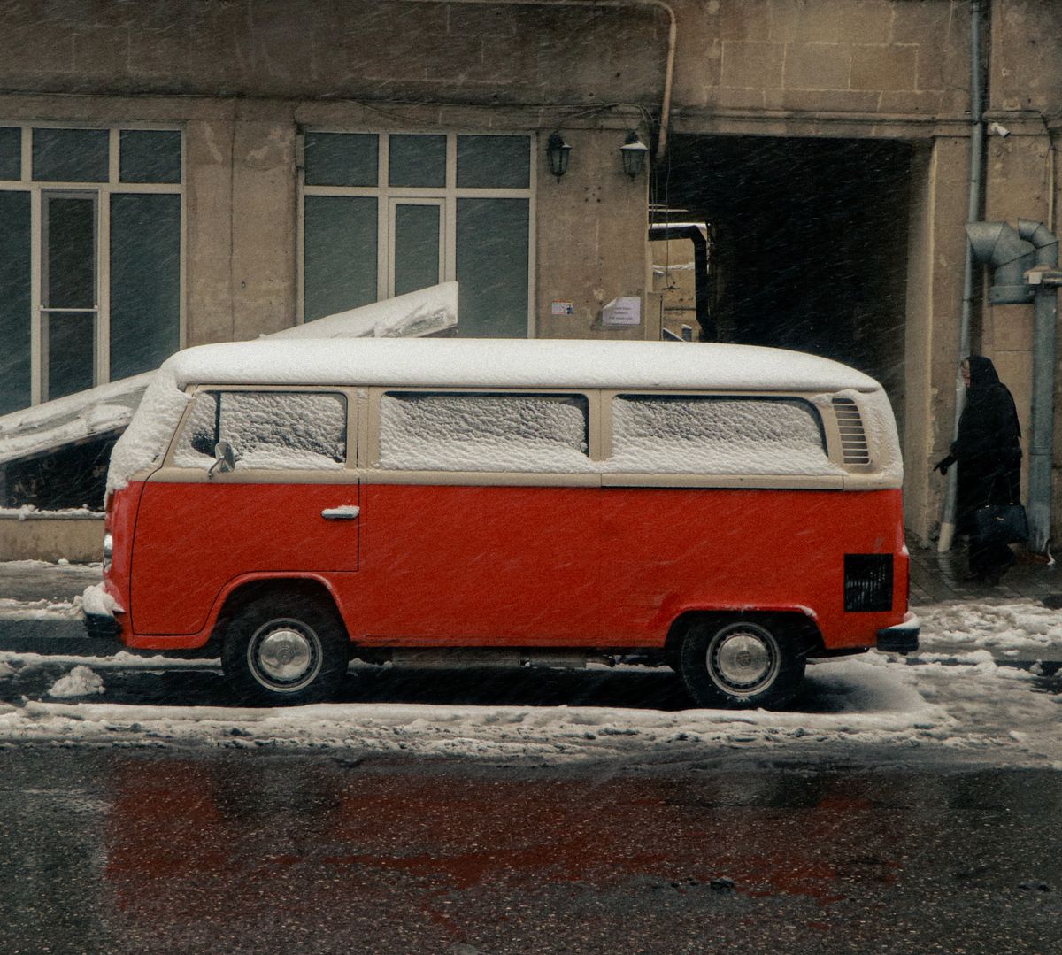 A vintage red bus covered in snow parked on an urban street during winter.