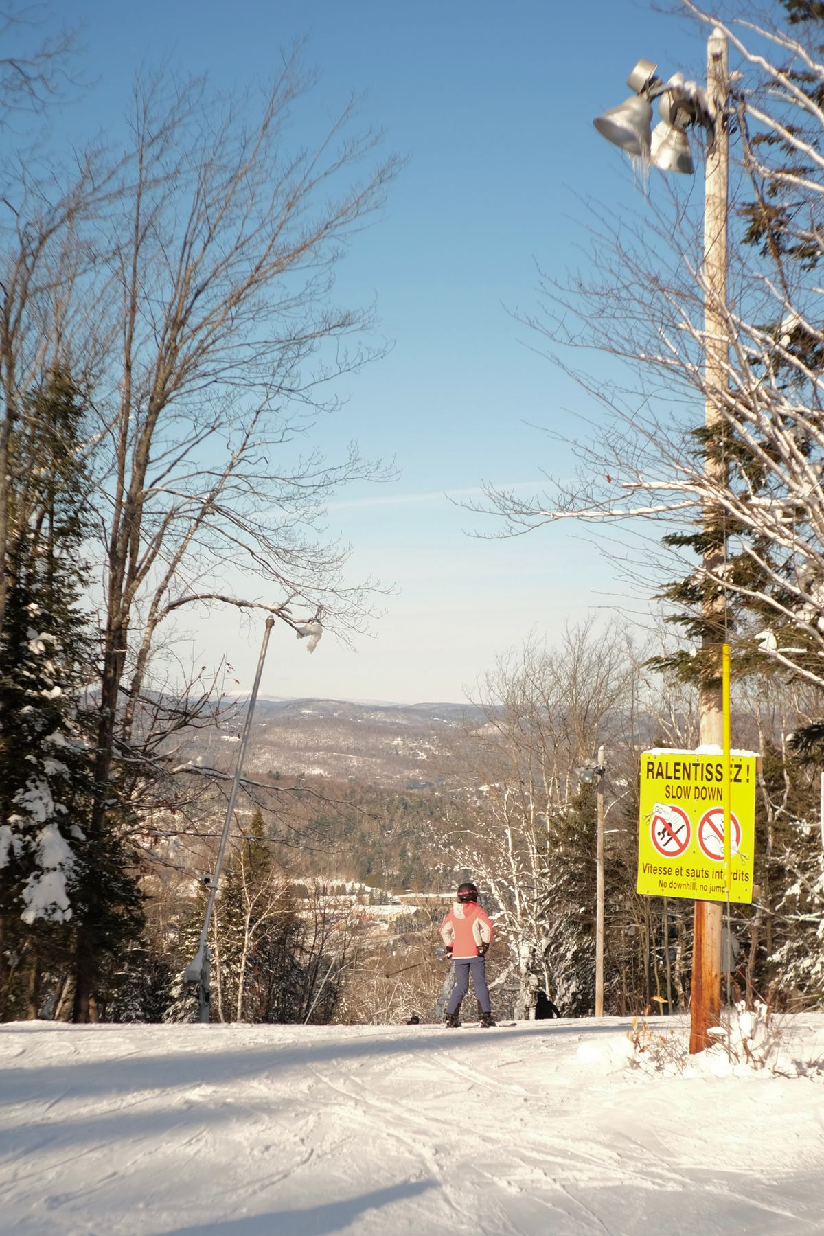 Skier on a snowy slope descending in a winter landscape, with a 'slow down' sign visible.