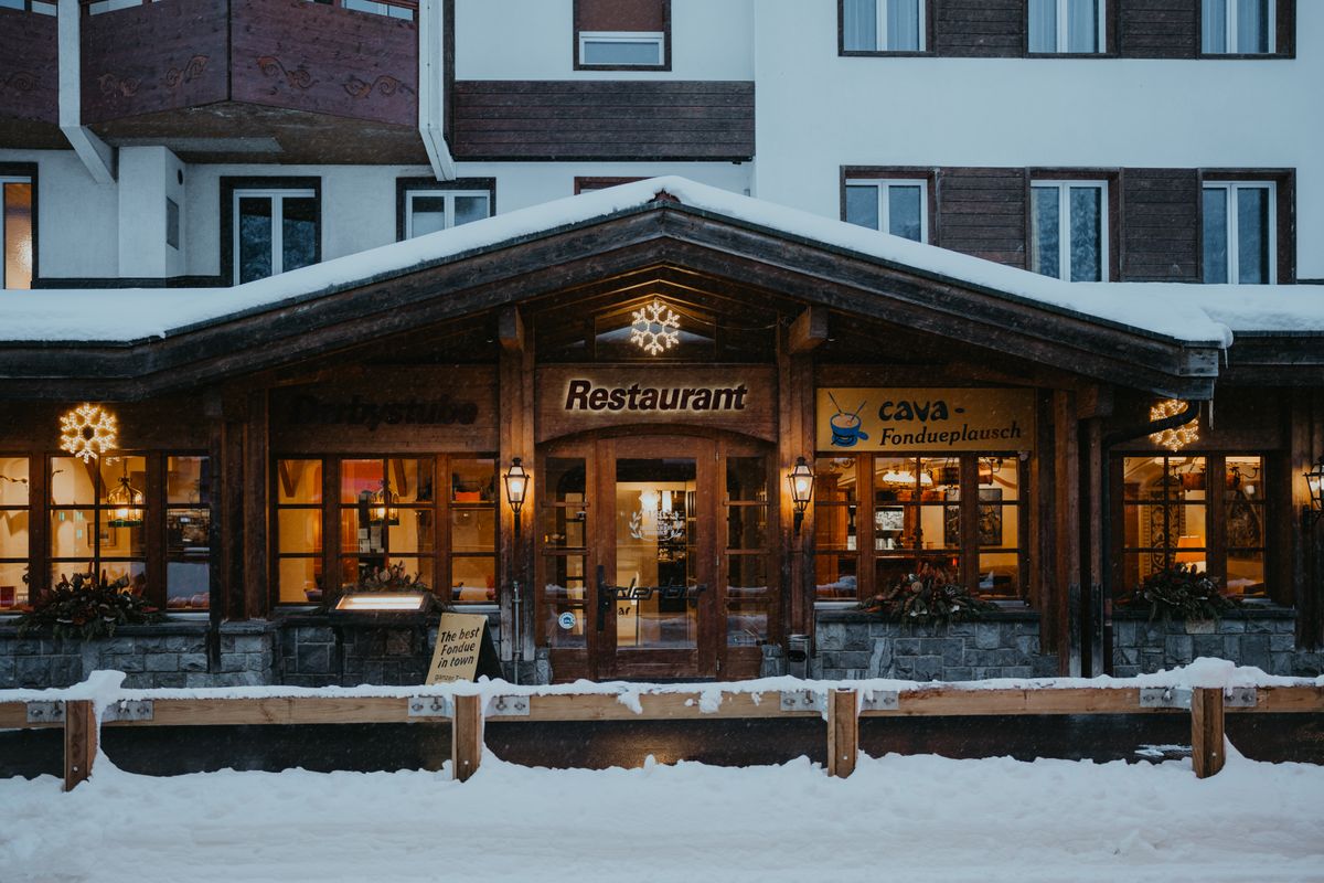 Cozy Swiss restaurant front with snow-covered surroundings and warm lights, inviting atmosphere.